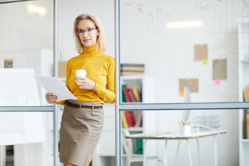 Blonde Frau im Business-Outfit lehnt an einer Glaswand und hält einen weißen Becher in der einen und Zettel in der anderen Hand