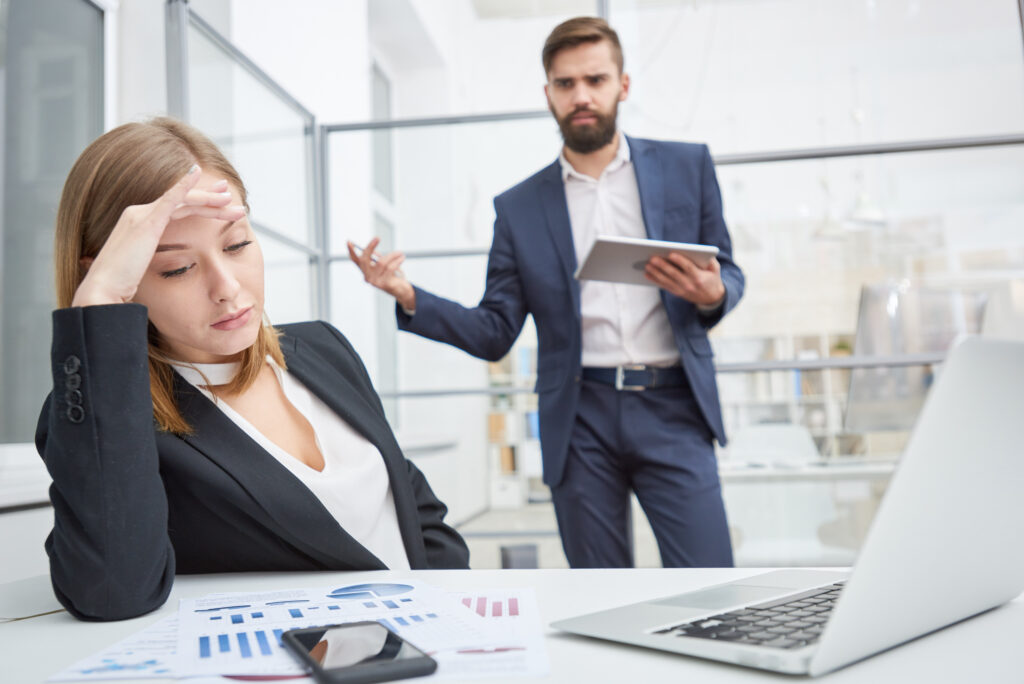 Frau in Business-Outfit blickt nachdenklich auf Unterlagen, Mann mit Anzug und Unterlagen in der Hand richtet Blick verärgert auf sie
