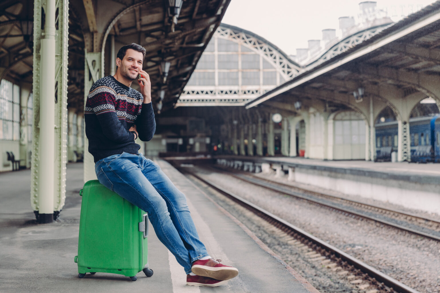Hübscher Mann mit Pullover und Jeans sitzt auf einem grünen Koffer, telefoniert und wartet auf den Zug