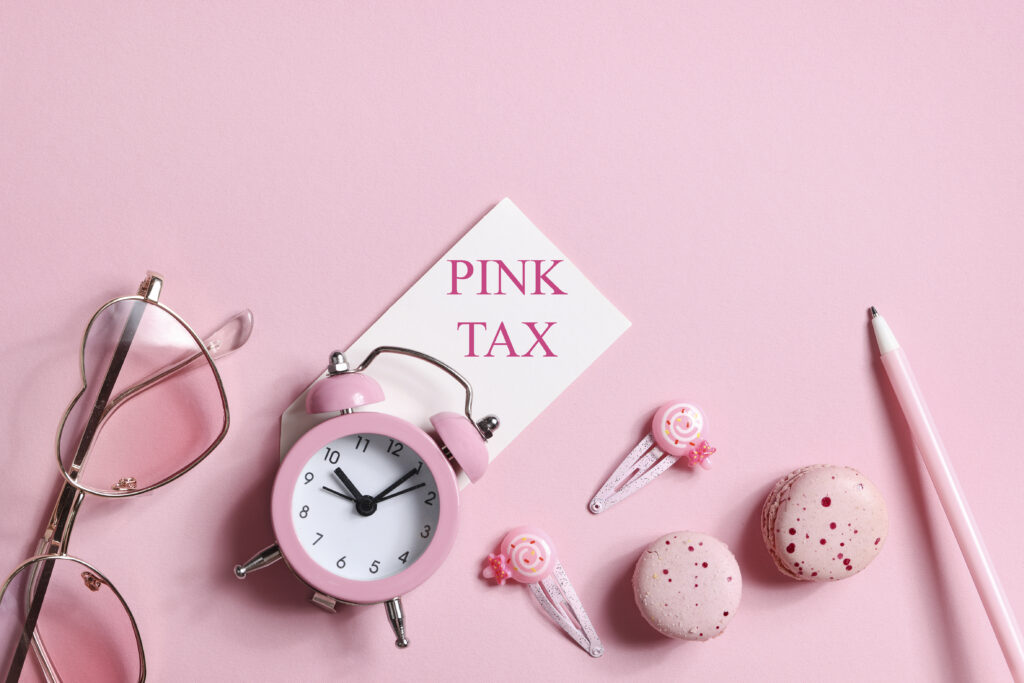 Rosaroter Hintergrund. Davor eine Brille mit Gläsern in Herzform, ein Wecker in rosa Design, die Worte Pink Tax auf einem weißen Schild, rosa Haarspangen, zwei Kekse und ein rosa Bleistift