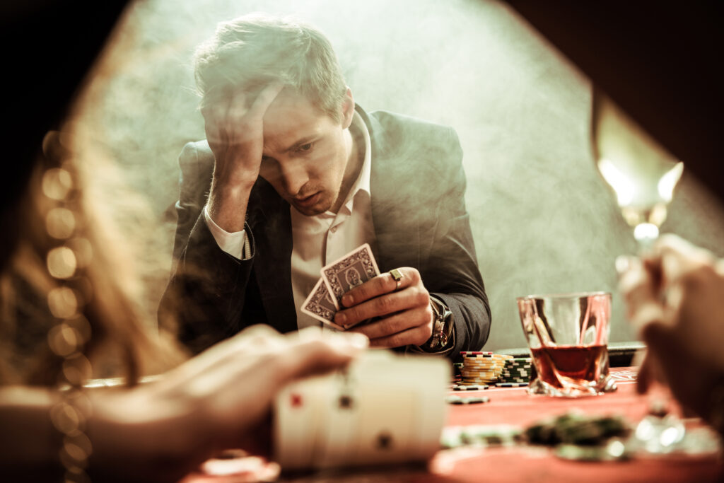 Ein Mann sitzt an einem Poker-Tisch mit zwei Karten in der Hand und greift mit mit der anderen Hand verzweifelt an die Stirn. 