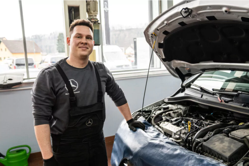 KFZ-Mechaniker steht in Arbeitsmontur neben einem Mercedes bei dem der Motordeckel geöffnet ist