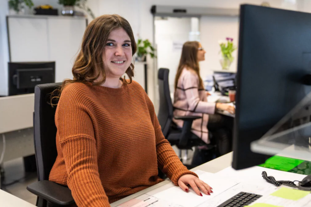 Frau in orangem Pulli sitzt an einem Schreibtisch vor einem PC und lacht in die Kamera