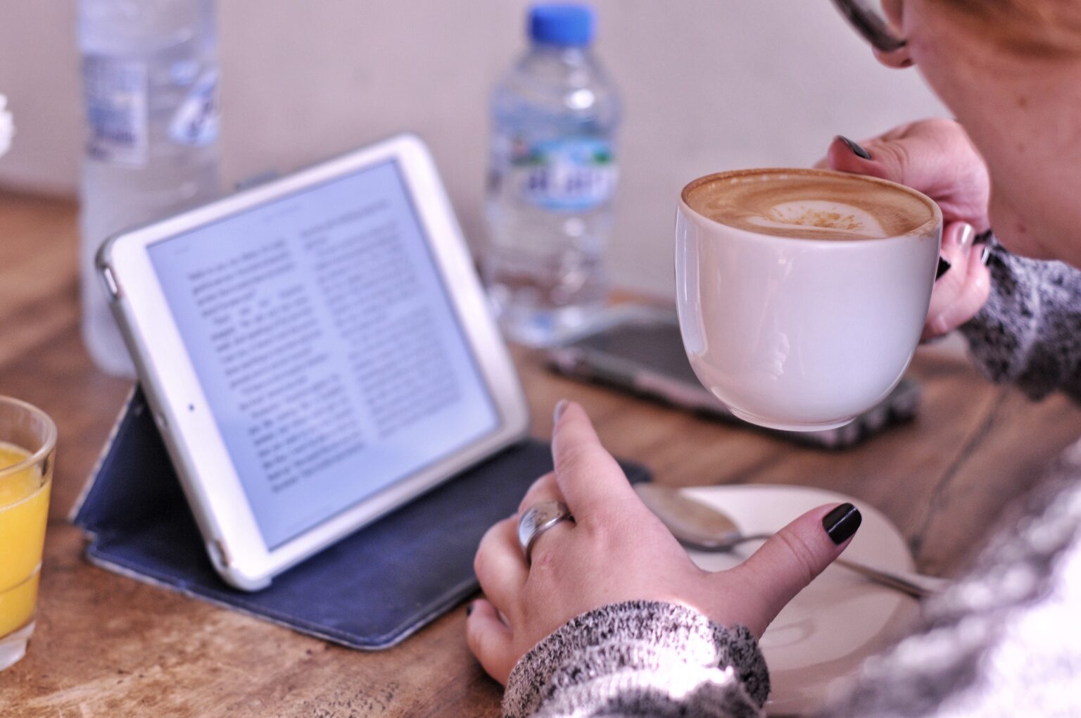 ebook auf einem elektronischen gerät auf einem Tisch. Davor eine Hand und eine andere Hand, die eine Kaffeetasse hält.