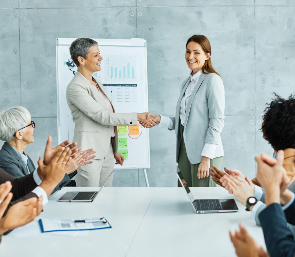 Zwei Frauen stehen vor einem Flipchart und reichen sich die Hand. Am Tisch sitzen vier Menschen die applaudieren