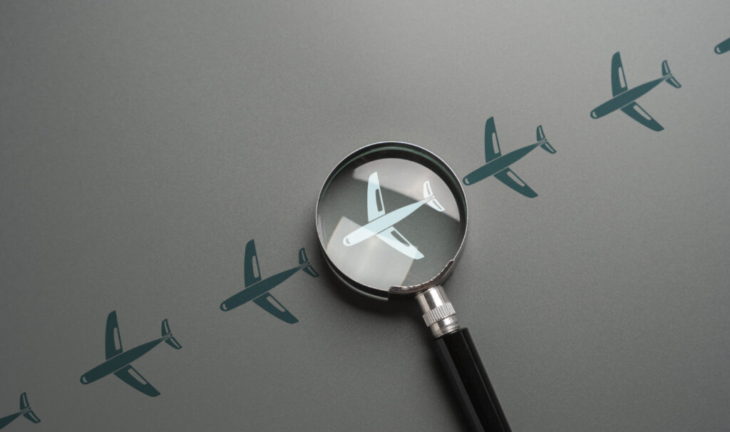 Grauer Hintergrund mit Zeichnungen von Flugzeugen. Darauf eine Lupe