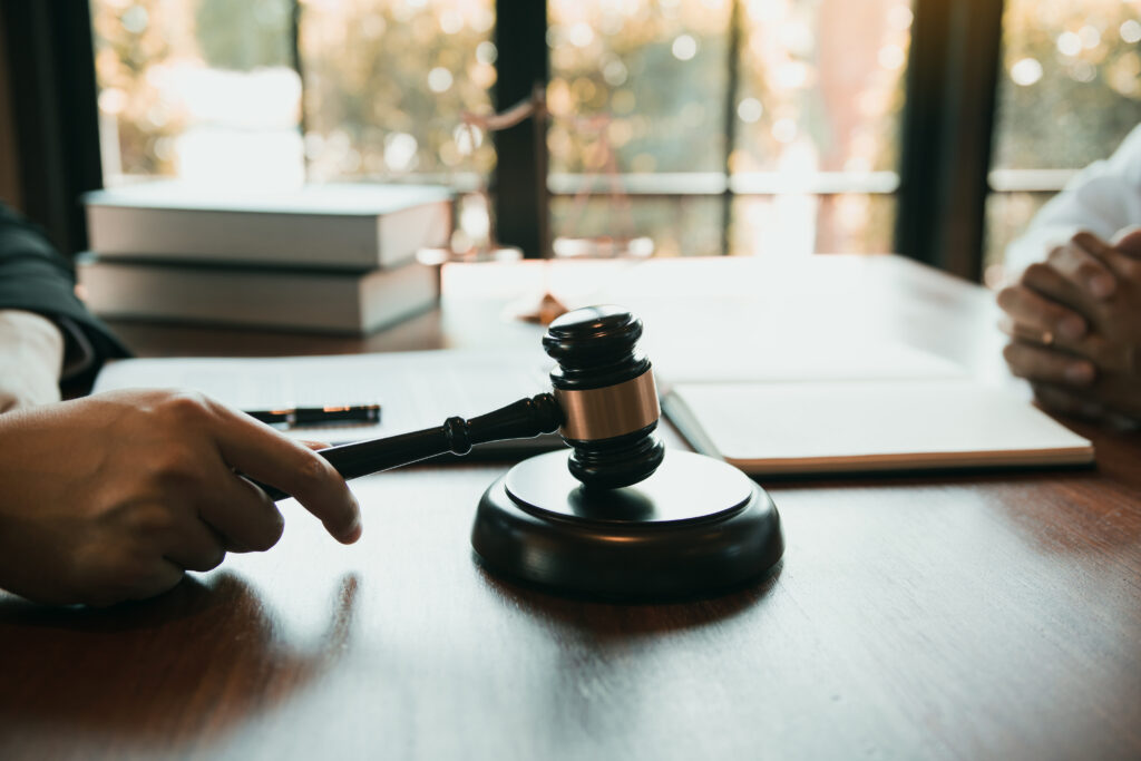 Tisch mit einer Hand, die einen Gerichtshammer auf den Klotz schlägt. Dahinter zwei Bücher und eine Schreibunterlage