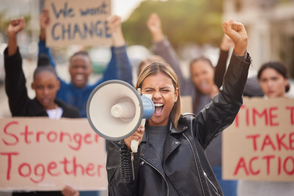 Frau bei einer Demonstration mit einem Lautsprecher in der einen Hand und die andere Hand zur Faust erhoben. Hinter hier einige Menschen mit Plakaten auf denen we want change, stronger togehter steht