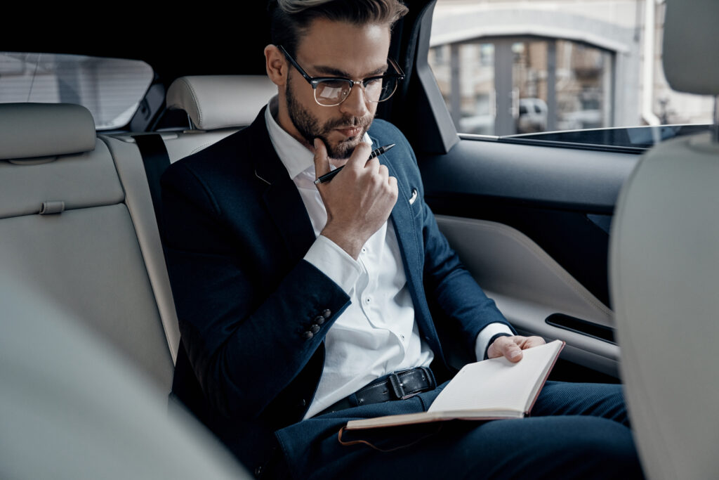 Businessman in dunklem Anzug und weißem Hemd sitzt in einem Auto. Er hält sich die Hand an sein Kinn. In der anderen Hand hält er eine Unterlage