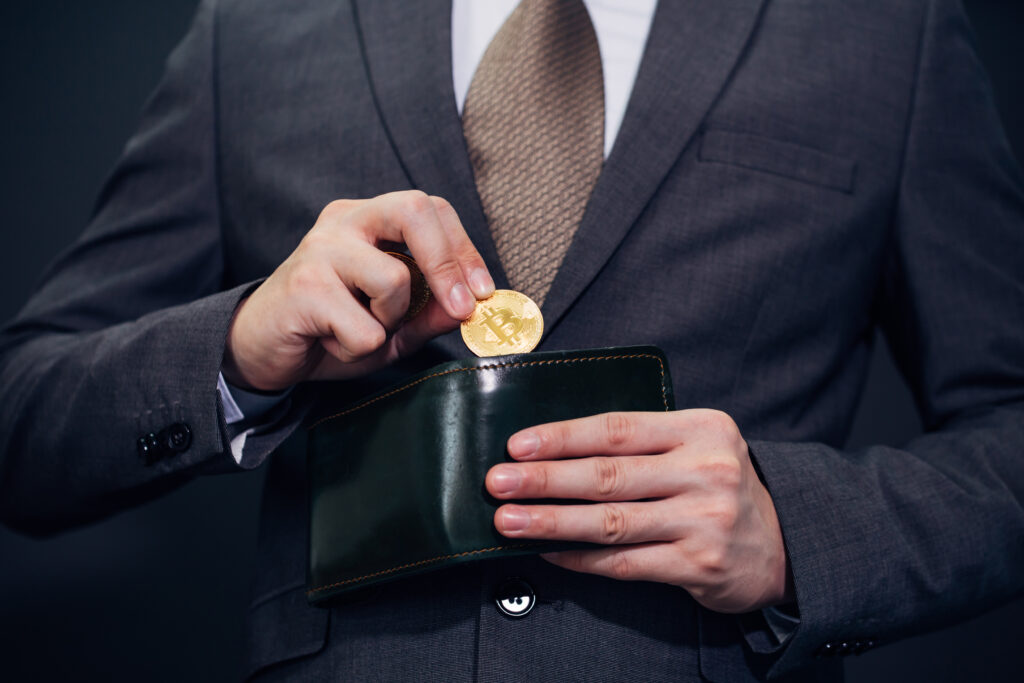 Mann in einem schwarzen Anzug hält einen Bitcoin und gib diesen in seine schwarze Geldtasche