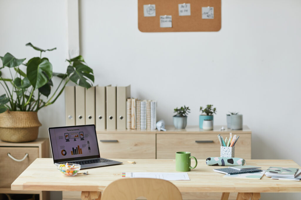 Home Office Holztisch mit einem PC, einem Kaffeehäferl in grün und verschiedenen weiteren Unterlagen. Davor ein Sessel, dahinter ein Holzregal auf dem Ordner, Bücher und ein paar Blumen stehen