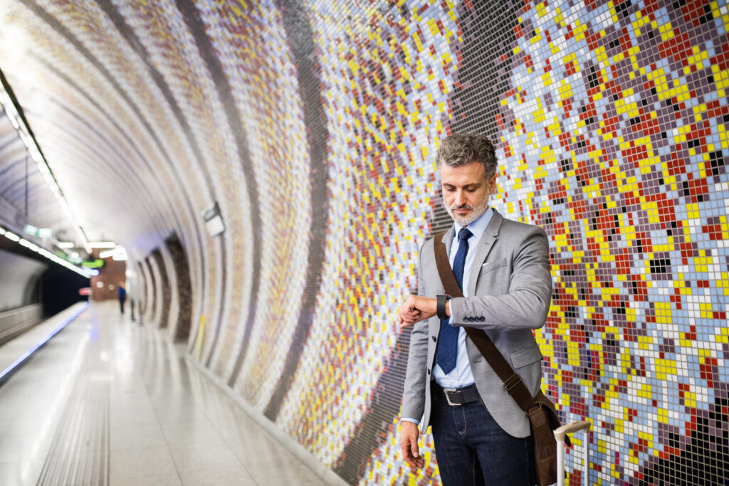 Mann mit hellem Sakko, Krawatte und umgehängter Tasche steht, blickt auf seine Uhr und wartet auf die U-Bahn