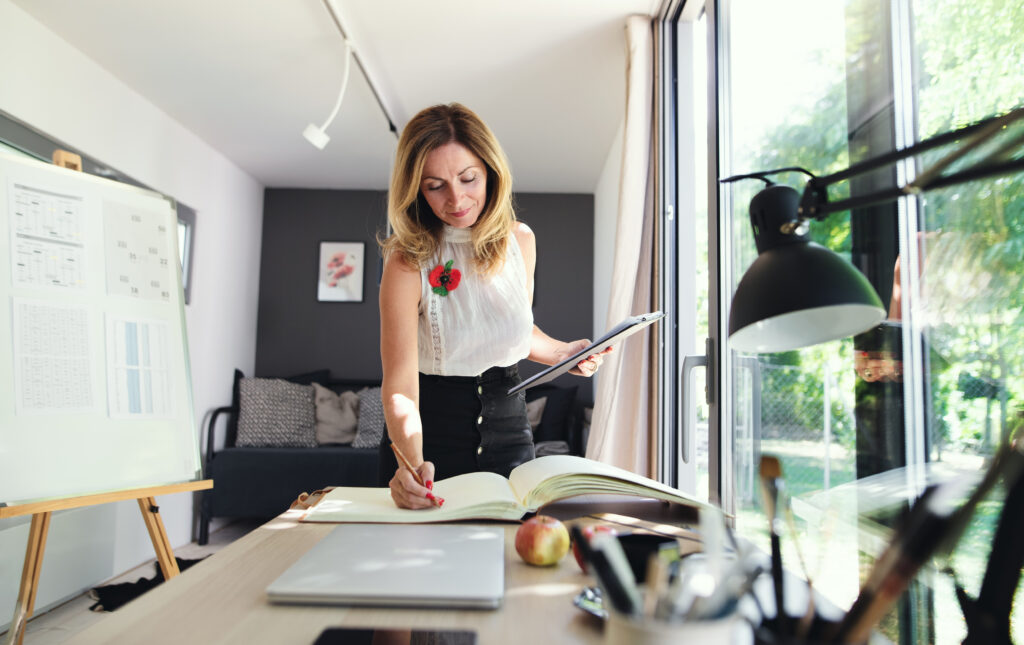 Home Office Fau mit einer weißen Bluse und einem schwarzen Rock steht an einem Tisch auf dem ein Buch liegt und macht Eintragungen. Neben ihr eine große Pinnwand und auf der anderen Seite eine große Glasfront