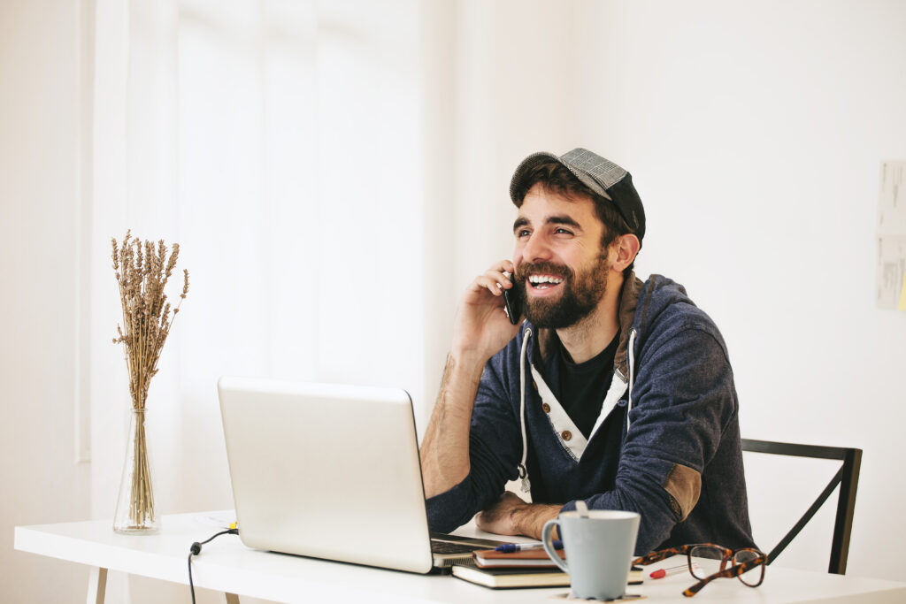 Home Office Mann mit Kappe am Kopf und blauem Sweat-Shirt-Pullover sitzt an einem Tisch mit einem PC, einer Kaffeetasse und einer Brille sowie Notzizbüchern. Er hält sein Handy an das Ohr