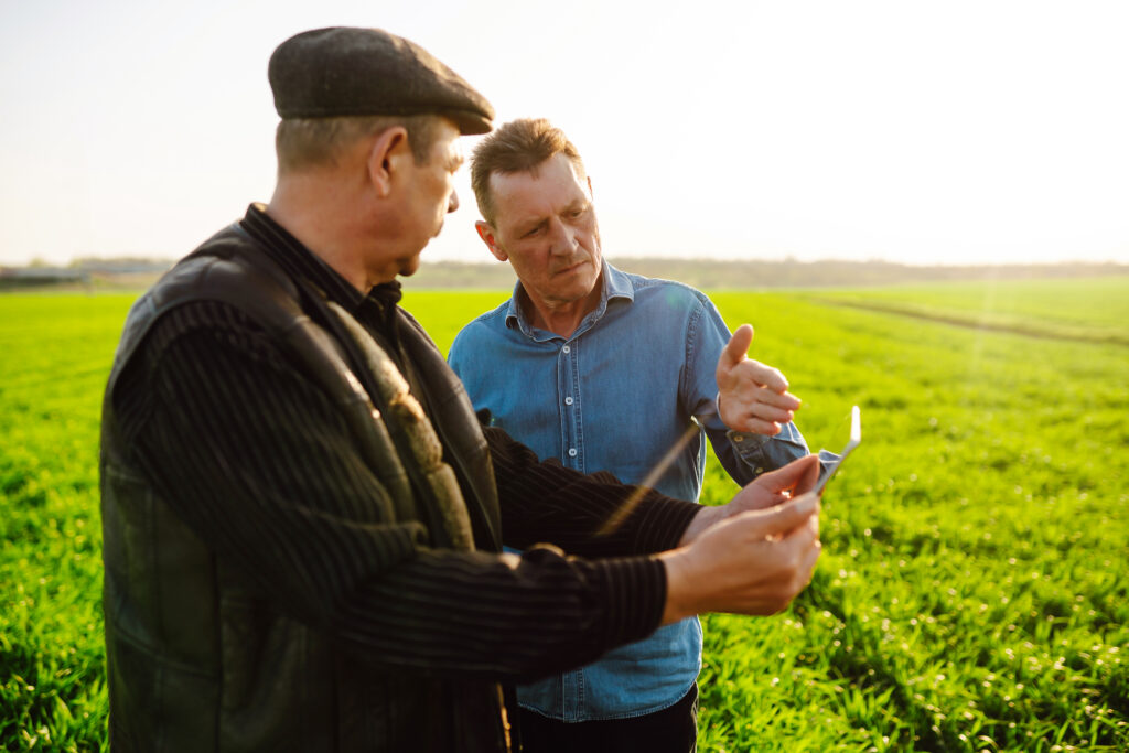 Smart Farming Zwei Männer stehen auf einem Feld. Der eine hält ein Tablett, der andere zeigt mit einer Hand darauf