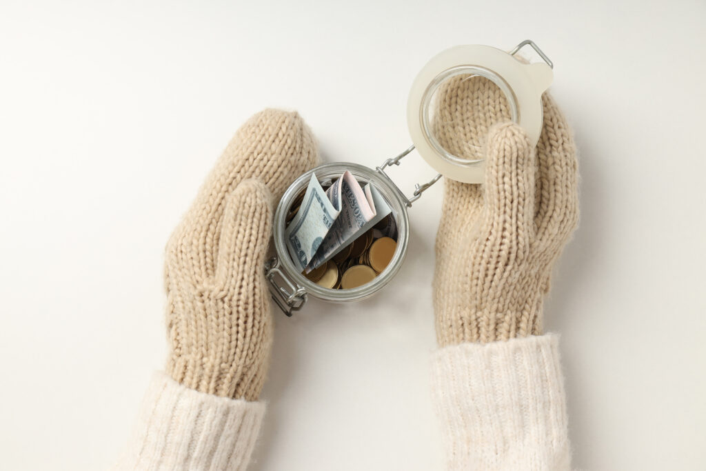 Hände mit weiße gestrickten Handschuhen die ein Glas halten indem ein paar Geldscheine und Münzen sind