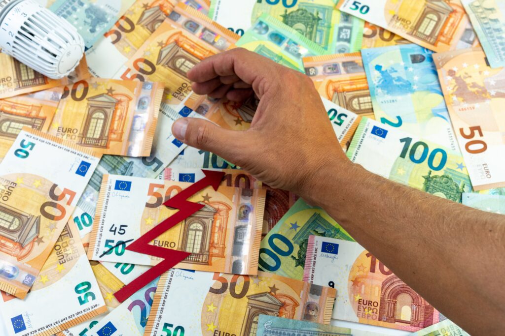 Viele Euro-Geldscheine, eine Hand, ein Heizregler und ein roter Pfeil der nach oben zeigt