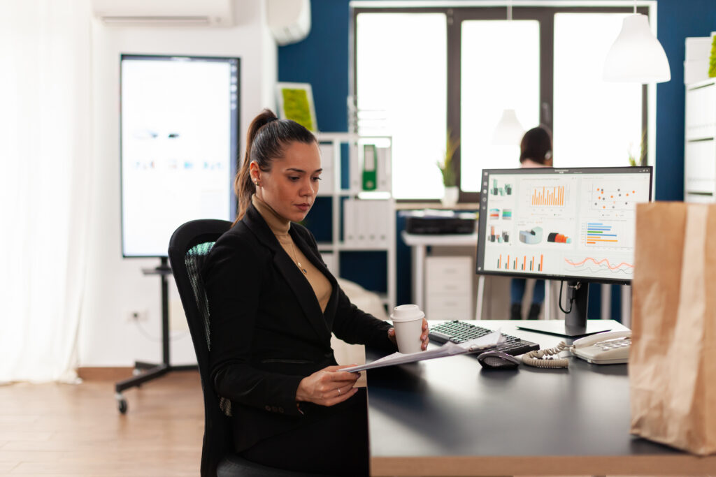 Frau hält einen Kaffee und eine Liste in ihren Händen. Sie sitzt an einem Schreibtisch mit einem PC 