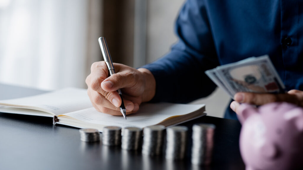 Hand hält einen Stift. Davor viele aufgestappelte Münzen