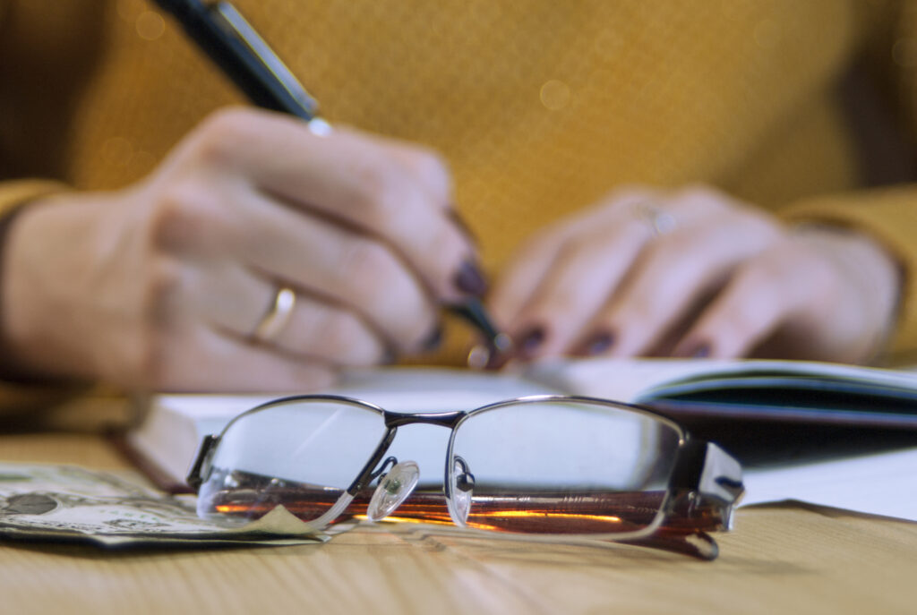 Nahaufnahme einer Brille. Dahinter eine Hand mit einem Stift die etwas in ein Buch schreibt