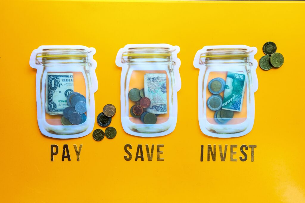 Drei leere Einmachgläser mit Pay, Save und Invest darunter geschrieben. In den Gläsern sind Münzen und Geldscheine
