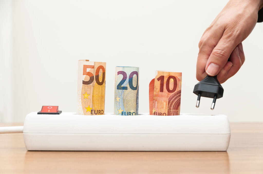 Stromsteckdose in der ein 50,20 und 10 Euroschein steckt und eine Hand einen Stecker in die letzte vierte Öffnung steckt