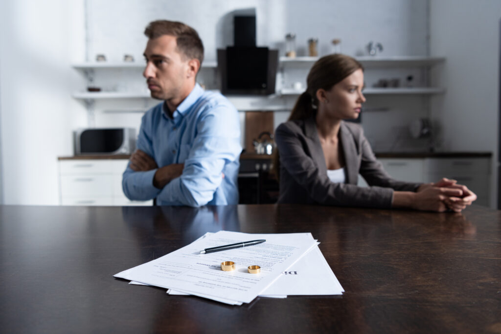 Mann und Frau sitzen mit dem Rücken zueinander an einem Tisch auf dem Papier und die Eheringe sowie ein Stift liegen